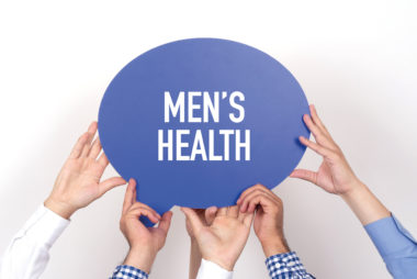 Hands of three men in blue, white, and plaid shirts holding up a blue speech bubble sign with the words “Men’s Health” in white text.