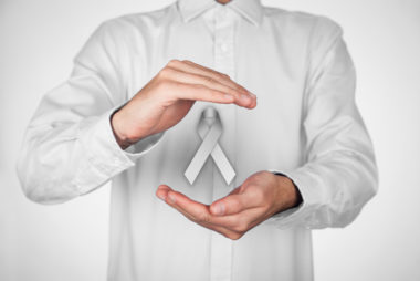 Close-up of man in off-white shirt with hands framing a floating grey Parkinson’s awareness ribbon on a white background.