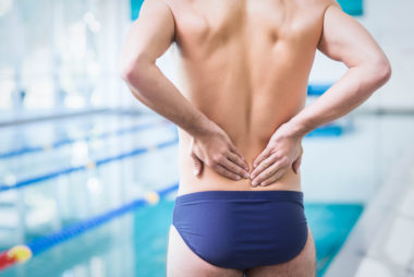 Fit man in swimwear standing poolside with both hands bracing his lower back, symbolising pain and stiffness associated with ankylosing spondylitis (AS).
