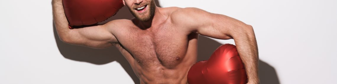 Smiling shirtless man with beard in black jeans holding two inflatable red heart balloons against a white background.
