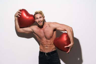 Smiling shirtless man with beard in black jeans holding two inflatable red heart balloons against a white background.