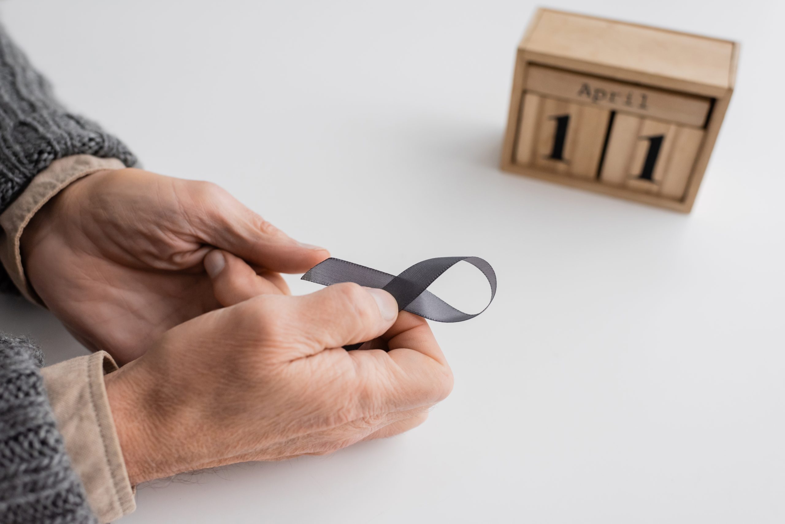 Senior man’s hands holding a grey awareness ribbon, with a blurred wood block calendar showing April 11 in the background, symbolising World Parkinson’s Day.