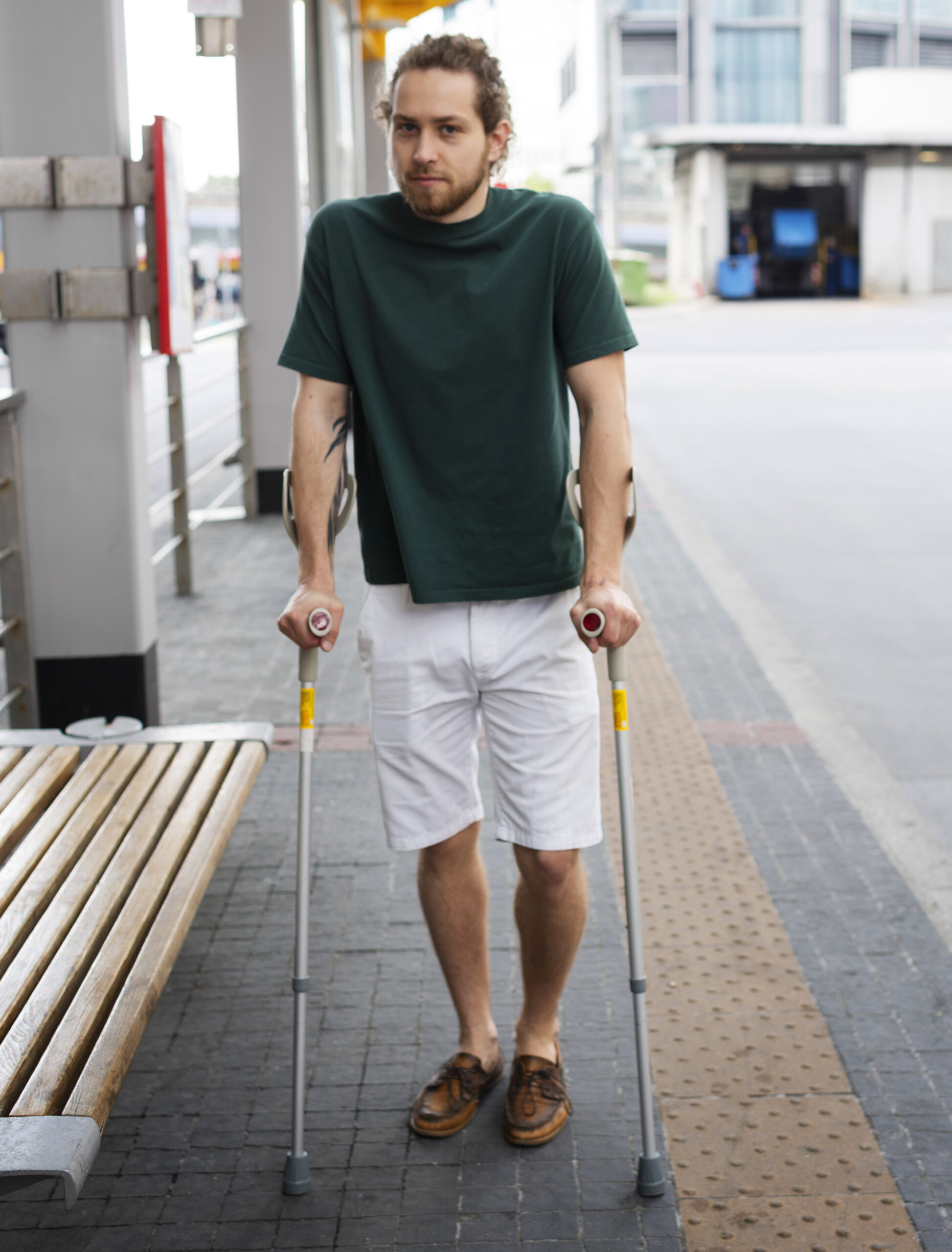 Young man with cerebral palsy using crutches at a city bus depot, with urban buildings and streetscape blurred in the background.