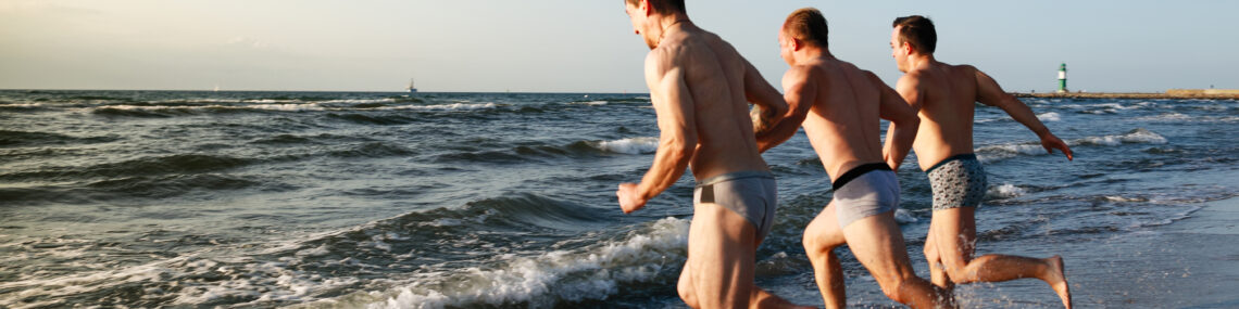 Three muscular men in underwear sprint into the ocean waves at the beach, splashing as they run side by side.
