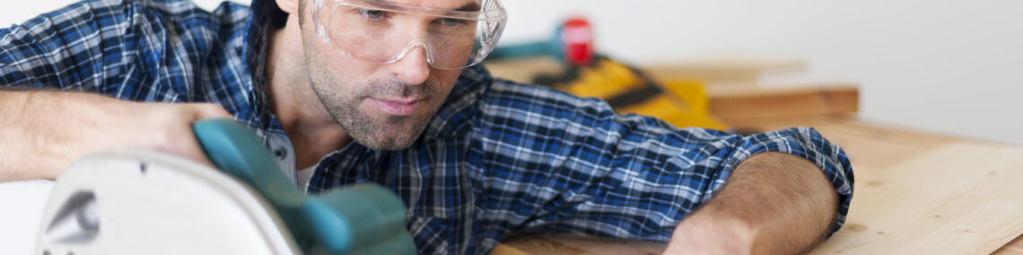 Close up of a carpenter in a blue plaid shirt on a work site, holding a circular saw in one hand and timber in the other with focused expression.