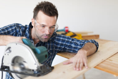 Close up of a carpenter in a blue plaid shirt on a work site, holding a circular saw in one hand and timber in the other with focused expression.