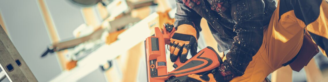 Carpenter wearing safety glasses and earmuffs using a nail gun on a building site, with timber truss and tools blurred in the background.