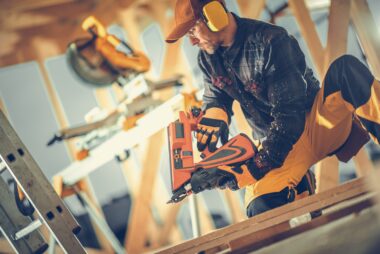 Carpenter wearing safety glasses and earmuffs using a nail gun on a building site, with timber truss and tools blurred in the background.