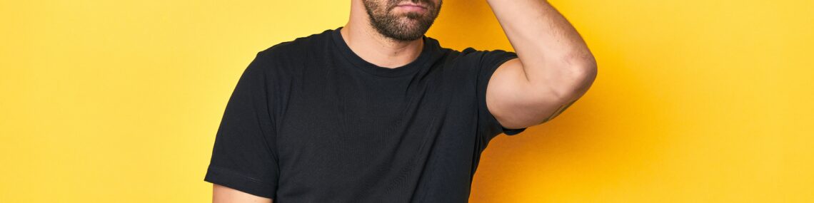 Man in black t-shirt against yellow background holding his head with a defeated expression, symbolising the emotional struggles highlighted on R U OK? Day.