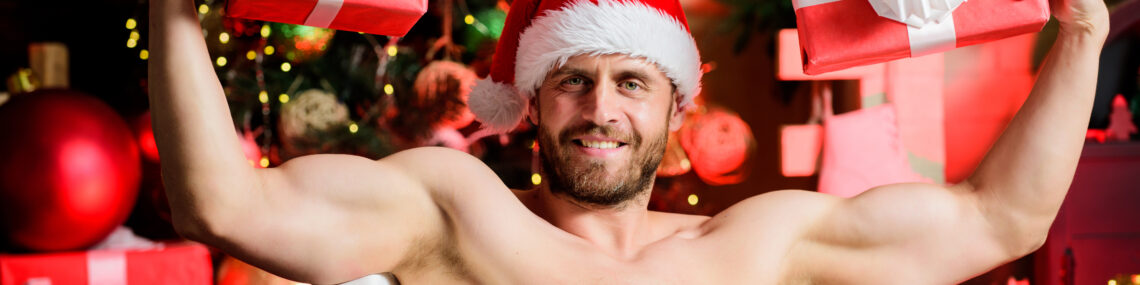 Muscular bearded man wearing a Santa hat, sitting naked in a bathtub and holding two wrapped Christmas presents, with a decorated tree and gifts blurred in the background.