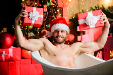Muscular bearded man wearing a Santa hat, sitting naked in a bathtub and holding two wrapped Christmas presents, with a decorated tree and gifts blurred in the background.