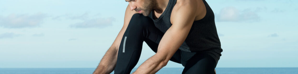 Close-up of a male runner in tights kneeling on one leg, holding his bare foot with concern after removing his shoe, on an asphalt path with ocean and blue sky in the background.
