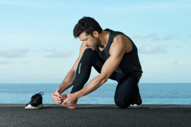 Close-up of a male runner in tights kneeling on one leg, holding his bare foot with concern after removing his shoe, on an asphalt path with ocean and blue sky in the background.