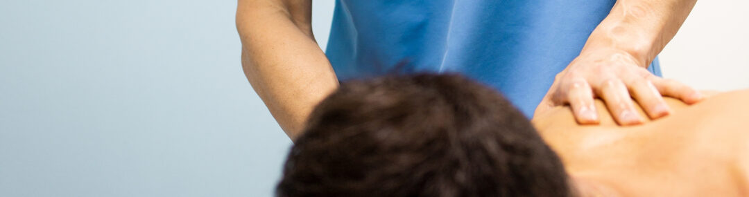 Clinical massage therapist in blue uniform treating a man lying face down on a treatment table.