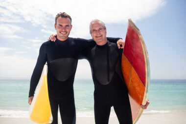 Portrait of a happy father and son in wetsuits embracing on the beach with surfboards, standing at the water’s edge after surfing together.