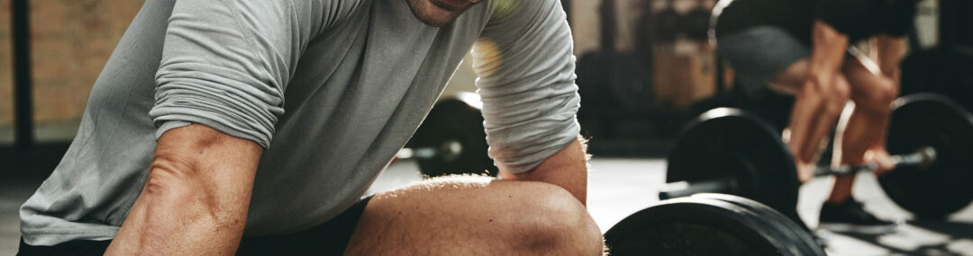 Two men lifting weights together in a gym, one in the foreground and the other out of focus in the background, showing camaraderie and strength.