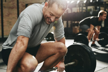 Two men lifting weights together in a gym, one in the foreground and the other out of focus in the background, showing camaraderie and strength.