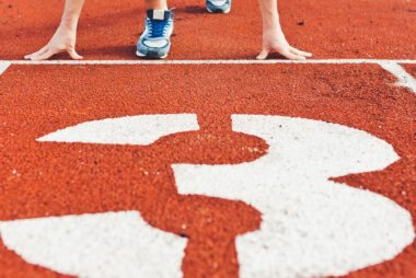 Man wearing blue running shoes in the start position on an athletic track in lane number 3.