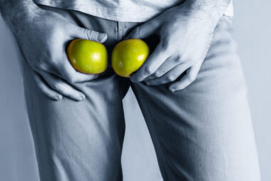Man holding two green apples over his groin, the only colour against an otherwise blue-tinted image to symbolise testicles.