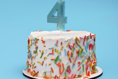 Number four birthday candle burning on a cake with heavy white frosting and multicoloured sprinkles against a light blue background.