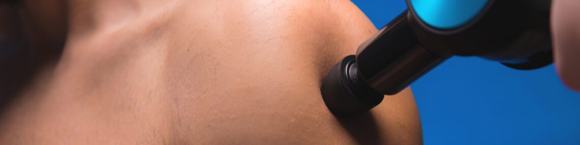 Close-up of a bearded man receiving percussion massage on his shoulder with a handheld device.