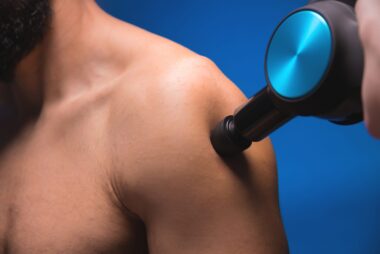 Close-up of a bearded man receiving percussion massage on his shoulder with a handheld device.