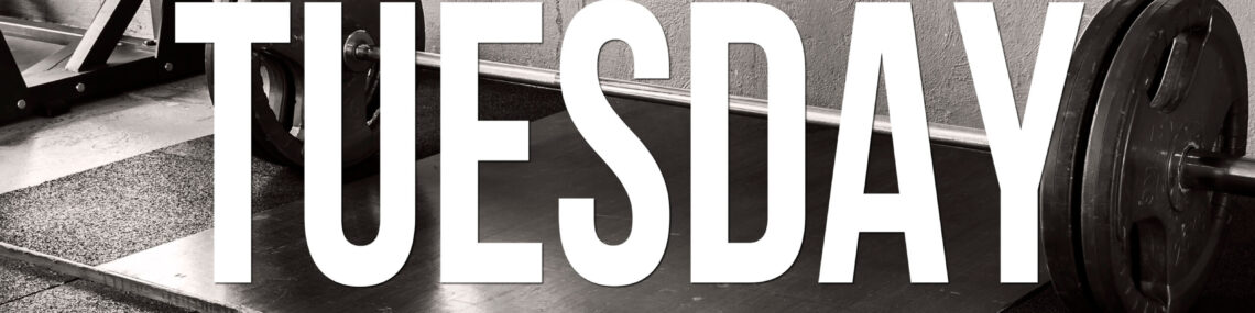 Black and white image of a barbell with weight plates resting on a gym floor, with the word TUESDAY in uppercase text overlaid on the image.