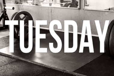 Black and white image of a barbell with weight plates resting on a gym floor, with the word TUESDAY in uppercase text overlaid on the image.