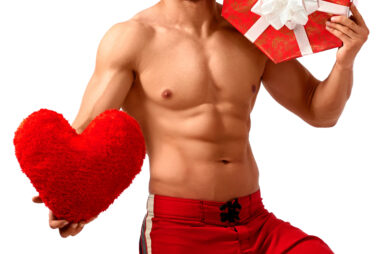 Shirtless man in red board shorts holding a plush heart-shaped cushion in one hand and a large red gift box with a white bow against his chest.