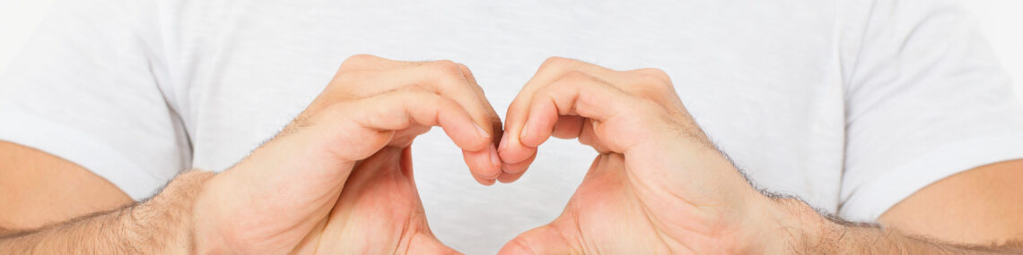 Close-up of a man in a white shirt making a heart gesture with his hands across his chest.