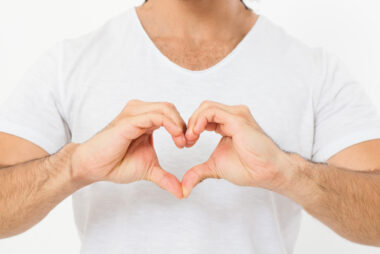 Close-up of a man in a white shirt making a heart gesture with his hands across his chest.