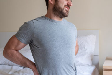 Man sitting on the edge of a bed holding his lower back in discomfort after sleeping.