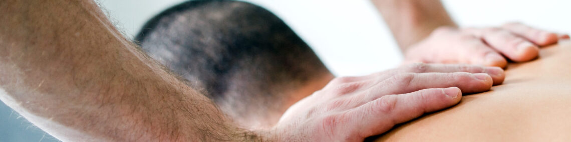 Man lying face down on a massage table in a clinical setting while a male therapist applies massage therapy to his shoulders.