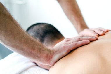 Man lying face down on a massage table in a clinical setting while a male therapist applies massage therapy to his shoulders.