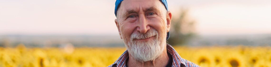Senior man wearing a backwards blue cap standing in a sunflower field at dusk, facing the audience.