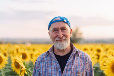 Senior man wearing a backwards blue cap standing in a sunflower field at dusk, facing the audience.